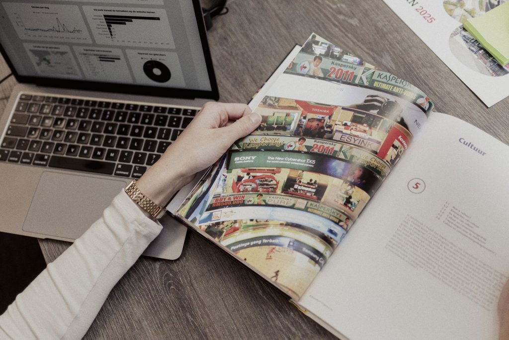 Een vrouw bladert door een kleurrijk magazine terwijl haar laptop open ligt met grafieken en dashboards in beeld.
