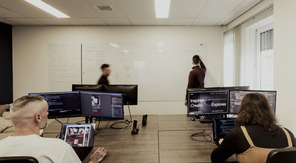 Twee mensen kijken naar twee grote computerschermen met code, terwijl een derde collega op een bord schrijft en iemand anders door de ruimte loopt