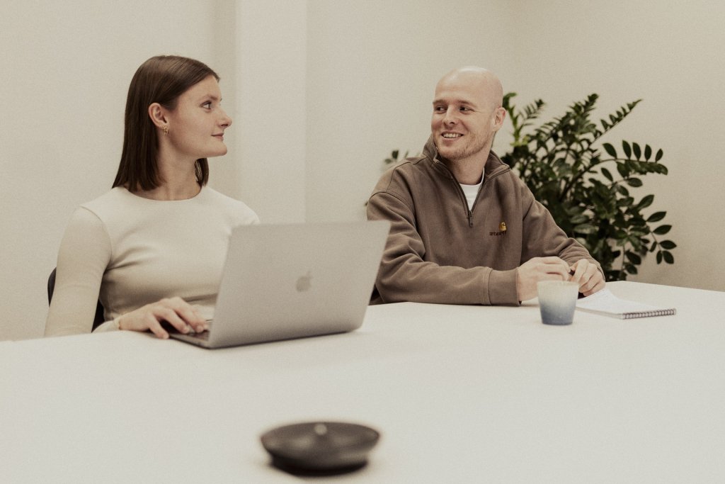 Een vrouw en een man zitten aan een tafel met een laptop tussen hen in en praten.